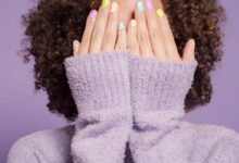 Mujer con suéter lila que se cubre la cara con las manos y muestra uñas pintadas en tonos pastel