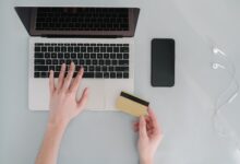 Manos sobre el teclado de una notebook con una tarjeta bancaria al lado del celular