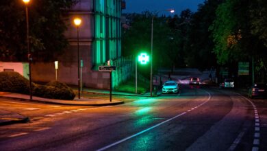 Cruz verde luminosa de una farmacia encendida de noche junto a una calle con faroles