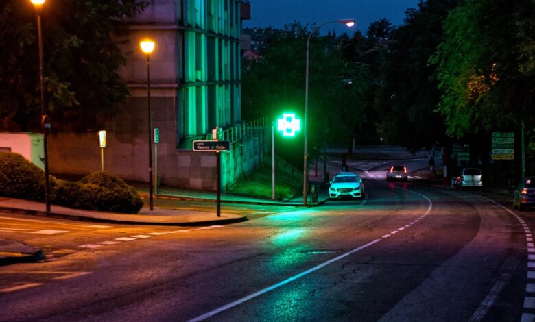 Cruz verde luminosa de una farmacia encendida de noche junto a una calle con faroles