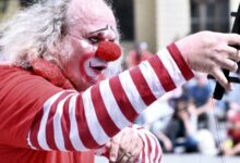 Payaso con nariz roja y traje a rayas actúa con un micrófono frente al público en la calle