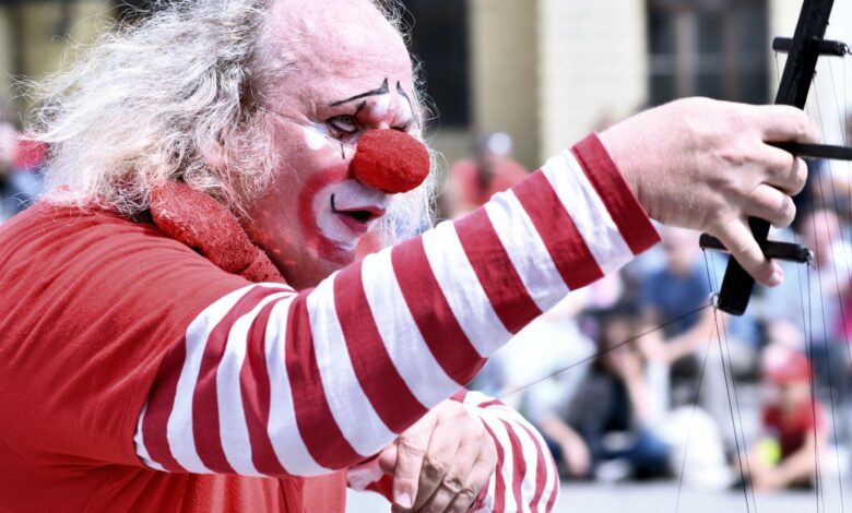 Payaso con nariz roja y traje a rayas actúa con un micrófono frente al público en la calle